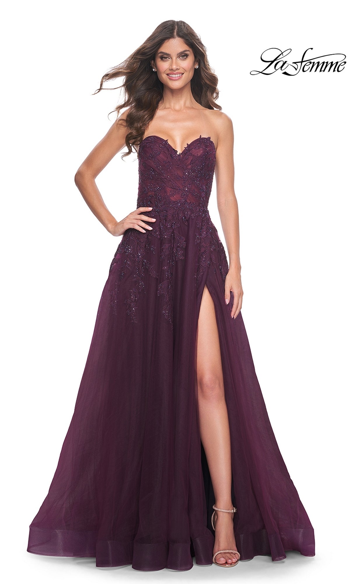 With a confident smile, a woman showcases the La Femme Lace-Up Long Sweetheart Prom Dress 32304, a deep purple strapless A-line gown featuring lace accents and a high slit for an elegant formal look.